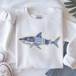May include: White sweatshirt with a shark design. The shark is made up of different blue patterns and textures, edged with a decorative trim. The sweatshirt has a crew neck and long sleeves, with the brand name "Gildan" visible on the tag.