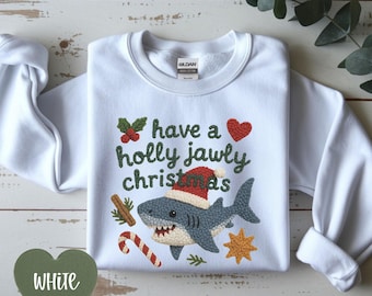 Puede incluir: Sudadera blanca con la frase "have a holly jawly christmas" y un tiburón con gorro de Papá Noel. El diseño incluye un corazón rojo, acebo, un bastón de caramelo y una estrella. La sudadera es de la marca Gildan.