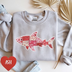 Op de afbeelding: Een lichtgrijs sweatshirt met een patchwork haaienontwerp. De haai is samengesteld uit verschillende rode en roze patronen, waaronder harten en lippen, met het woord "LOVE" op het ontwerp. Een rood hart met het woord "ASH" staat in de linker benedenhoek.