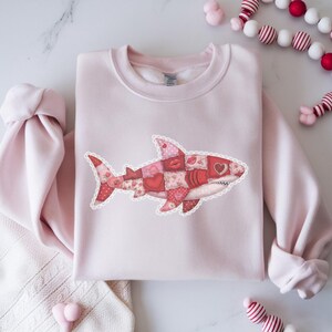 May include: A pale pink sweatshirt with a patchwork shark design. The shark is decorated with hearts, lips, and floral patterns in red and pink. It has a white, scalloped border. The sweatshirt is neatly folded on a white surface.