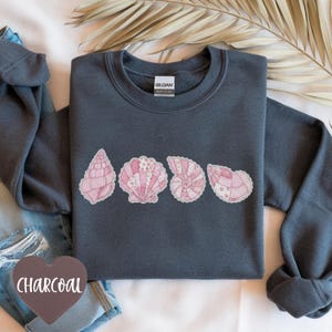 May include: A charcoal gray sweatshirt featuring a row of five pink seashell designs. The seashells have intricate patterns and textures. The sweatshirt has long sleeves and a crew neck. The word "CHARCOAL" is on a heart-shaped tag.