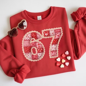 Pode incluir: Um moletom vermelho com o número "67" aplicado na frente em um design de patchwork de corações rosa e vermelhos. Óculos de sol dourados em forma de coração e doces em forma de coração também são visíveis.