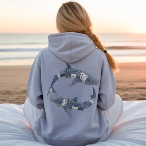 May include: A light gray hoodie featuring a design of two whale sharks in a circular pattern on the back. The whale sharks have a blue and white patchwork design. The hoodie has a hood and long sleeves.