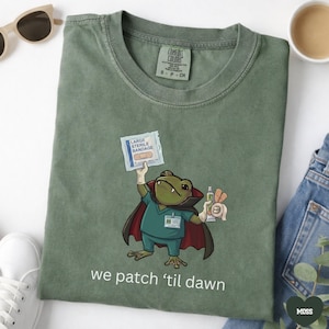 May include: A moss green t-shirt featuring a graphic of a frog dressed as a vampire doctor wearing a teal scrub top and a dark cape. The frog holds a large sterile bandage and medical supplies. The text reads, we patch ‘til dawn.