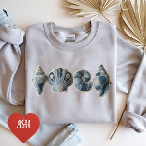 May include: A light gray crewneck sweatshirt featuring a graphic print of four distinct seashells arranged in a row. Each shell displays a blue and white patchwork-style pattern with intricate details. The brand label reads Gildan.