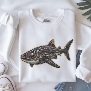 Puede incluir: Una sudadera blanca de cuello redondo con un gráfico de un tiburón ballena relleno de ilustraciones detalladas de Halloween que incluyen calaveras, huesos, fantasmas, una bruja con un caldero y gatos negros, en tonos morados y grises.