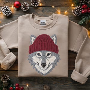 May include: A tan sweatshirt featuring an embroidered wolf wearing a burgundy knit cap. The wolf has gray and white fur, blue eyes, and a black nose. The sweatshirt has long sleeves and a crew neck.