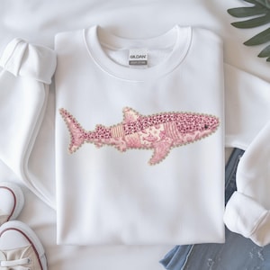 May include: White sweatshirt featuring a pink shark design. The shark is composed of various patterns, including coral, leopard print, and stripes. The design is outlined with a scalloped edge. The sweatshirt has a crew neck and long sleeves.