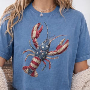 Puede incluir: Una camiseta azul de manga corta con un gran gráfico de una langosta. El diseño de la langosta está compuesto por parches de tela rojos, blancos y azules, con un patrón de estrellas blancas en la cabeza y rayas en la cola y las pinzas.