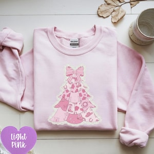 Könnte beinhalten: Hellrosa Sweatshirt mit einem rosa Weihnachtsbaum-Design. Der Baum besteht aus verschiedenen rosa Mustern, darunter Kuhfellmuster, Stiefel und ein Hufeisen. Das Design wird von einer gestreiften Schleife gekrönt. Das Sweatshirt ist von der Marke Gildan.