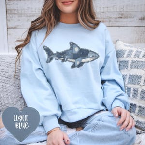 May include: Pale blue sweatshirt with a shark design made from denim patches. The shark is the focal point on the front. A heart-shaped tag in the lower left corner reads "LIGHT BLUE". The model is wearing blue jeans.