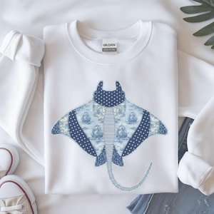 May include: A white crewneck sweatshirt featuring a large graphic of a manta ray. The manta ray design is composed of various blue and white patterned fabrics, including a nautical scene with sailing ships and a dark blue fabric with white polka dots.
