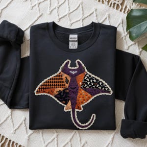May include: A black long-sleeve sweatshirt featuring a large, central graphic of a manta ray. The manta ray is composed of various patchwork-style patterns in orange, purple, and black, including motifs of bats, ghosts, pumpkins, and skulls.