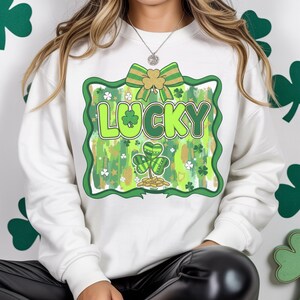 May include: White sweatshirt with a St. Patrick's Day design. The design features the word "LUCKY" in green, a green shamrock, a gold bow, and gold coins. The design is framed in green with a gold and green background.
