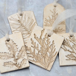 Könnte beinhalten: Fünf handgefertigte Geschenkanhänger aus Keramik mit rustikalem, natürlichem Look. Jeder Anhänger zeigt ein anderes geprägtes Design eines Blatt-Zweigs in hellbrauner Farbe. Die Anhänger sind mit einem weißen Band gebunden.