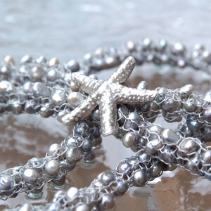 K&ouml;nnte beinhalten: Eine silberne Kette mit einem silbernen Seestern-Anh&auml;nger und grauen Perlen. Die Kette ist aus Draht und Perlen gefertigt.