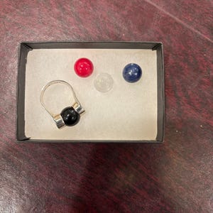 May include: A silver ring with a black bead and three small, colorful beads: red, clear, and blue. The ring and beads are displayed in a black box with white padding, set against a dark wood-grain surface.