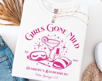 Puede incluir: Camiseta blanca con un gráfico rosa que dice "Girls Gone Mild" con un cóctel, antifaz, esmalte de uñas y zapatillas. El texto "Charlotte's Bachelorette, Palm Springs, CA" también está impreso.
