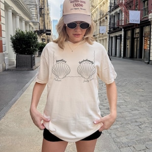 Puede incluir: Camiseta color crema con diseño de conchas marinas y el texto "Amelia's Bachelorette Tulum July 2025." La persona lleva una gorra beige con el texto "Western House Casino Las Vegas Nevada" y gafas de sol.