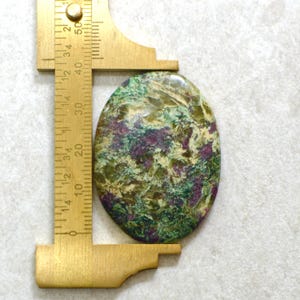 以下が含まれることがあります： 緑、紫、クリーム色の混合色を持つ楕円形の宝石。 金色の定規が石の隣に配置され、センチメートル単位の測定値が表示されています。 石の表面はテクスチャ加工されています。
