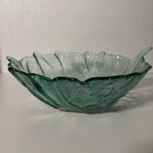 Op de afbeelding: Een heldere, getextureerde glazen kom met een bladachtig ontwerp. De kom heeft een blauwgroene tint en een golvende, geschulpte rand. De kom wordt waarschijnlijk gebruikt om voedsel in te serveren of als decoratief stuk.