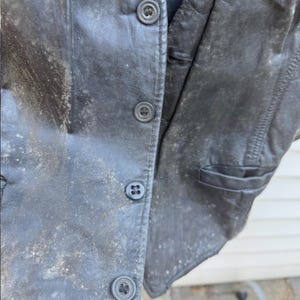 May include: Close-up of a weathered black leather vest with three button closures. The vest shows signs of wear and age, with a faded appearance and visible texture. The vest has a pocket on the right side.