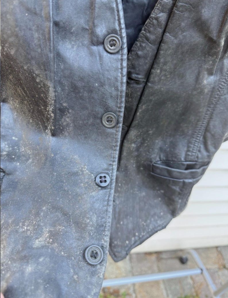May include: Close-up of a weathered black leather vest with three button closures. The vest shows signs of wear and age, with a faded appearance and visible texture. The vest has a pocket on the right side.