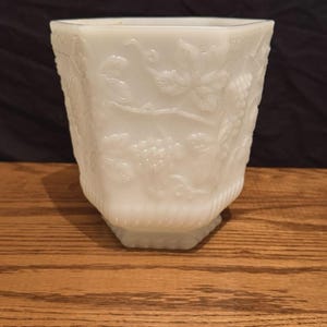 May include: A vintage, off-white glass vase with a hexagonal shape. The vase features a detailed embossed design of grape vines and leaves. The vase sits on a wooden surface.