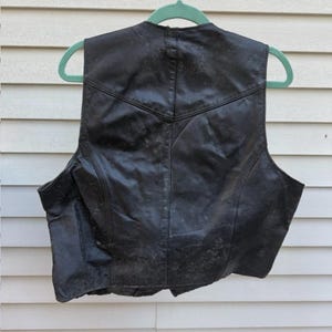 May include: Black leather vest with a classic design, hanging on a green hanger. The vest has a V-neck and a tailored fit, with visible stitching details. The leather appears worn, adding character to the garment.