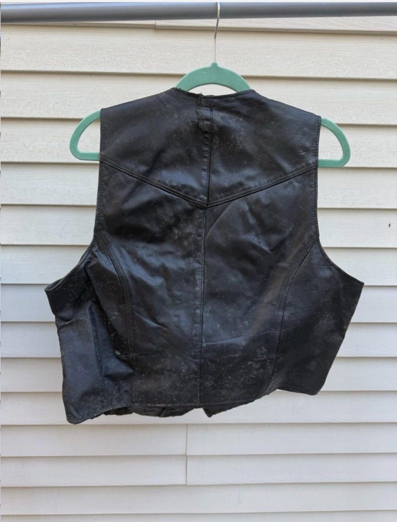 May include: Black leather vest with a classic design, hanging on a green hanger. The vest has a V-neck and a tailored fit, with visible stitching details. The leather appears worn, adding character to the garment.