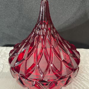 May include: A ruby red, lidded glass jar with a geometric diamond pattern. The jar has a rounded base and a conical, pointed top. The glass is transparent, allowing light to pass through and highlight the intricate design.