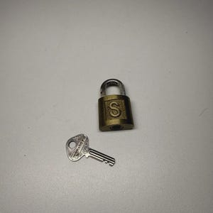 May include: A small, brass-colored padlock with the letter "S" on the front, next to a silver-colored key. The key is lying on a light gray surface. The padlock has a curved shackle.