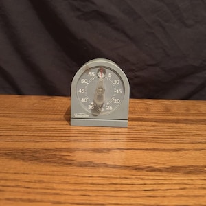 May include: A gray Sunbeam kitchen timer with a semi-circular top and a flat base. The face features a circular dial with black numbers from 5 to 55, a central clear plastic knob, and a thin red indicator needle pointing toward the top position.