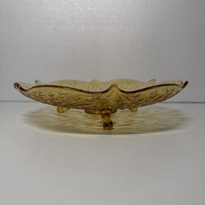 May include: A vintage amber-colored glass serving dish. The dish has a scalloped edge and is supported by three small feet. The interior of the dish features a delicate leaf pattern. This decorative piece is ideal for serving snacks or displaying small items.