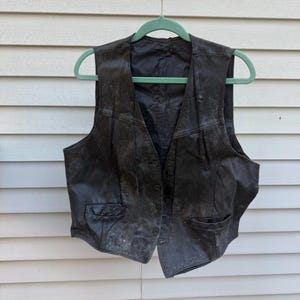 May include: A black leather vest with a button-up front and two front pockets hangs from a green hanger. The vest appears worn, with a weathered texture and visible wear marks. The vest is a classic piece of apparel.