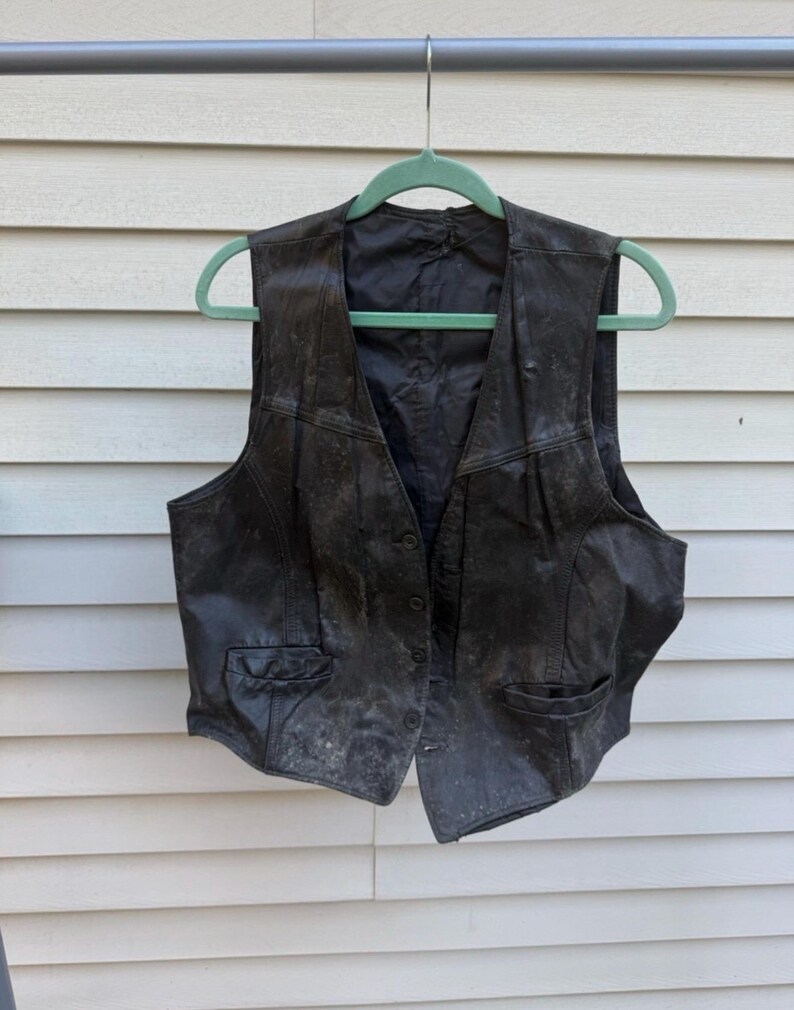 May include: A black leather vest with a button-up front and two front pockets hangs from a green hanger. The vest appears worn, with a weathered texture and visible wear marks. The vest is a classic piece of apparel.