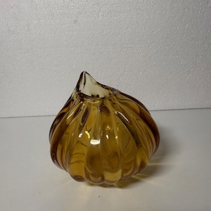 May include: Amber-colored glass decorative object with a bulbous shape and a pointed top. The glass has a ribbed texture, creating a play of light and shadow. The object is likely a decorative vase or bowl.