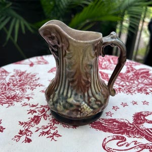 May include: A small ceramic pitcher featuring a glossy glaze in shades of brown, green, and tan. The exterior surface is decorated with a raised, vertical leaf and vine pattern, a curved handle, and a prominent pouring spout at the top rim.