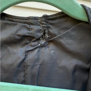 May include: Close-up of the inside of a black garment, possibly a jacket or coat, with visible damage to the lining near the collar. A green hanger is partially visible at the top and bottom of the frame.