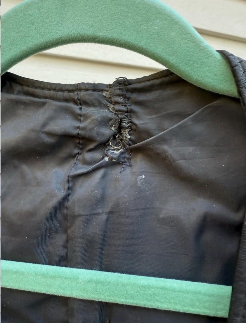 May include: Close-up of the inside of a black garment, possibly a jacket or coat, with visible damage to the lining near the collar. A green hanger is partially visible at the top and bottom of the frame.