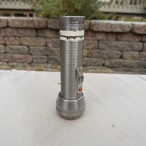 May include: A vintage silver flashlight with a ribbed body and a white band near the top. The flashlight has a rounded base and a switch with an orange button. The background is a brick wall and foliage.