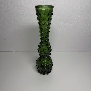 May include: A green glass vase with a textured, hobnail design. The vase has a bulbous base and a slender, elongated neck. The dark green colour and unique texture make it a decorative piece for home decor.
