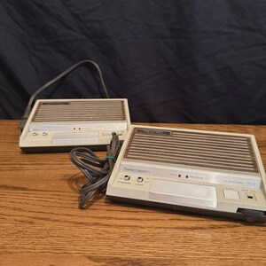 May include: Two beige and brown Realistic Selectacom 2-channel FM intercom units. Each device features a horizontal speaker grille, a silver control panel with buttons labeled Talk, Receive, Channel, Lock, and Call, and an attached power cord.