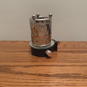 May include: A plug-in wax warmer featuring a cylindrical silver-tone metal sleeve with a decorative lattice cutout pattern. The top rim is accented with three small silver spheres. It has a black base with a silver control knob and a wall plug.