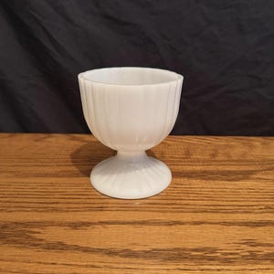 May include: A white, ribbed egg cup with a pedestal base. The egg cup is made of opaque glass and sits on a wooden surface. The background is a dark, solid color.