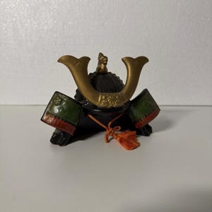 Puede incluir: Miniatura de casco de samurái japonés con cuernos y detalles dorados. El casco presenta paneles laterales verdes y rojos y una base negra. Una borla naranja cuelga de la parte delantera, añadiendo un toque decorativo.