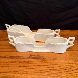 May include: Two white ceramic sleigh-shaped serving dishes with decorative floral patterns. Each dish has a handle on each end. The dishes are on a wooden surface.