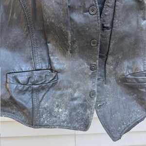 May include: A worn black leather vest with button closures and two front pockets. The vest shows signs of wear and tear, with visible discoloration and spots across the surface. The vest is a vintage garment.