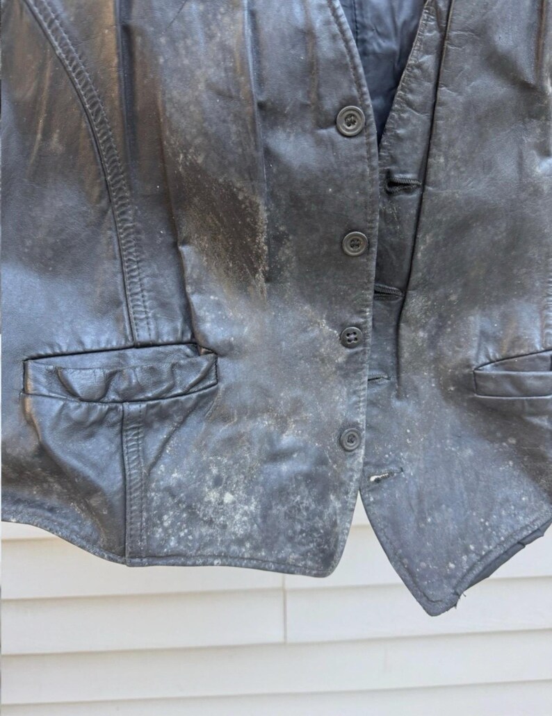 May include: A worn black leather vest with button closures and two front pockets. The vest shows signs of wear and tear, with visible discoloration and spots across the surface. The vest is a vintage garment.