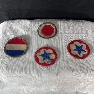 May include: A collection of vintage military patches. The patches feature various designs, including a red circle, a star within a flower shape, and a design with red, white, and blue stripes. They are set against a white patterned background.